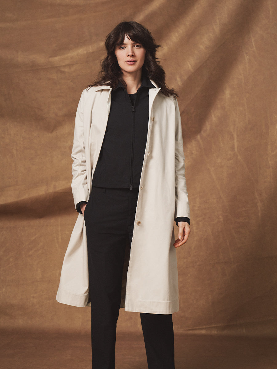 Woman wearing a beige trench coat over a black outfit against a brown background